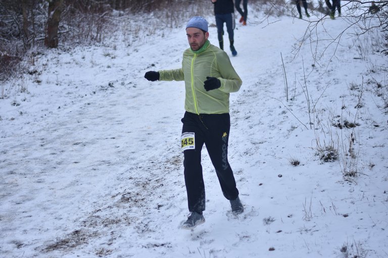 Muž běží po zasněžené cestě, má na sobě světle zelenou bundu, černé kalhoty se startovním číslem 145, černé rukavice a modrou čepici. V pozadí se objevují další běžci, holé stromy a zasněžená zem.