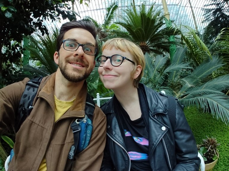 Dva lidé pózují na selfie v bujném skleníku plném hustých tropických rostlin a síťovaného stropu. Osoba nalevo má na sobě hnědou bundu přes žlutou košili a nese modrý batoh. Osoba napravo má na sobě černou koženou bundu přes černé tričko s barevnými vzory a černý batoh. Oba nosí brýle a pozadí je plné zelené listoví.