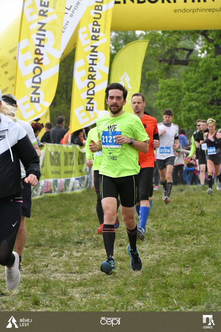 Skupina běžců soutěží na travnaté cestě během závodu. Centrální běžec má na sobě jasně zelené tričko se startovním číslem 2121, černé kraťasy a modré boty. Žluté bannery s nápisem „Endorphin Republic“ a odpovídající oblouk označují trať. Stromy a diváci lemují pozadí, s logy události („Běhej Lesy“, „ČEPS“) viditelnými dole. Jedná se o sponzorovanou běžeckou událost, nikoli bikepacking.