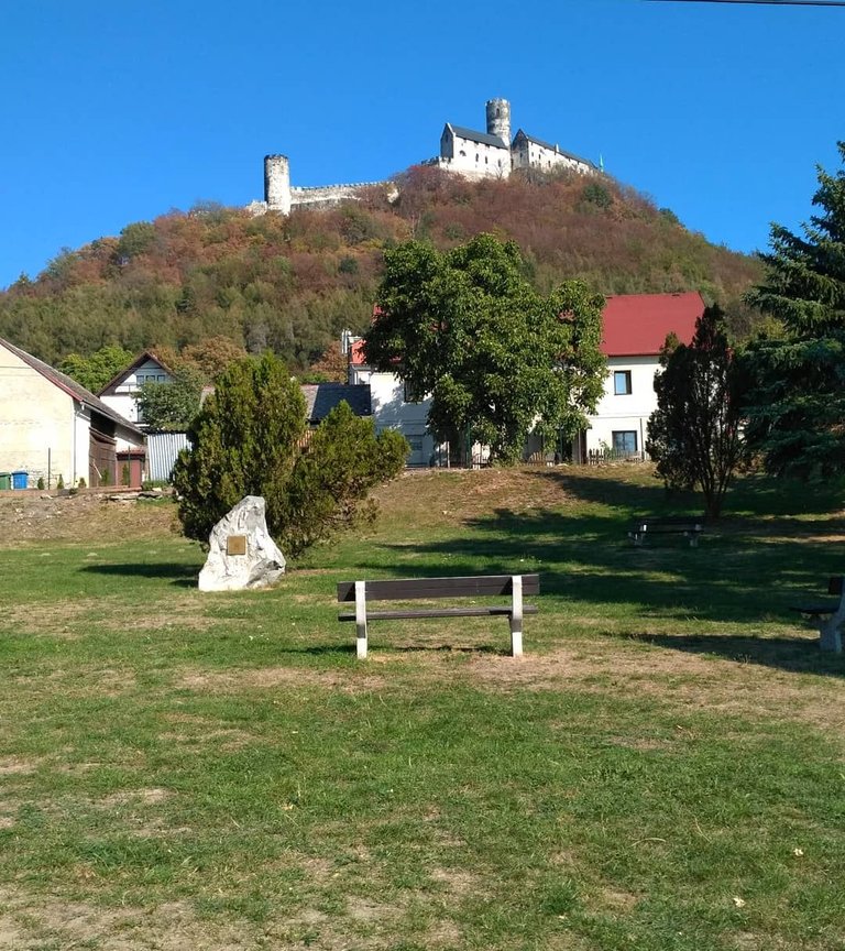 V popředí se nachází travnaté pole se dvěma dřevěnými lavičkami a velkým kamenným památníkem. Za nimi se rozprostírají domy s červenými střechami a roztroušené stromy, které vedou k podzimně zbarvenému kopci. Na vrcholu kopce stojí středověký hrad s kamennými věžemi, tyčící se proti jasně modré obloze – pravděpodobně malebná zastávka na bikepackingové trase.
