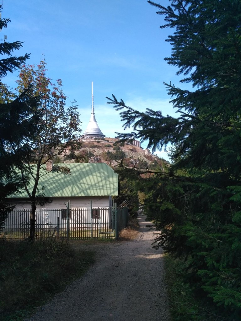 Štěrková cesta se vine mezi jehličnatými stromy směrem k bílému domu se zelenou střechou, ohraničenému kovovým plotem. Za domem se tyčí skalnatý kopec, na jehož vrcholu stojí vysoká, bílá, kuželovitá telekomunikační věž pod jasně modrou oblohou. Scéna naznačuje venkovskou nebo horskou cyklistickou trasu.