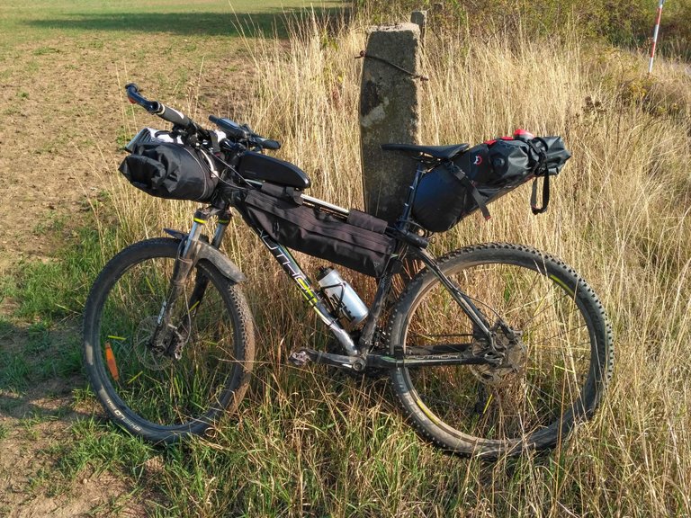 Černý horské kolo naložené černými bikepackingovými brašnami (na rámu, řídítkách a sedle) se opírá o betonový sloup v travnatém poli se suchou vegetací a hliněnou cestou. Kolo má tlusté pláště, kotoučové brzdy a láhev na vodu a nachází se ve venkovské přírodní oblasti.