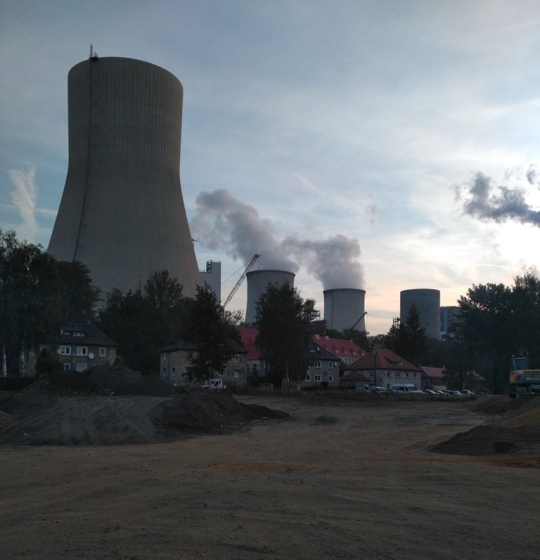Průmyslový areál za soumraku s velkými betonovými chladicími věžemi (některé vypouštějí kouř), staveništěm s hromadami hlíny v popředí, budovami s červenými střechami, stromy a jeřáby. Nejedná se o scénu z bikepackingu.