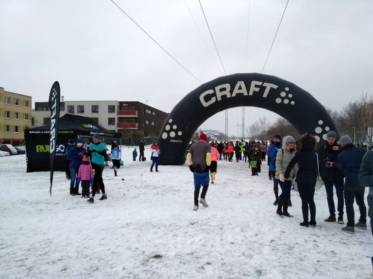 Zasněžená venkovní scéna s velkým černým nafukovacím obloukem, na kterém je bílými písmeny nápis „CRAFT“. Lidé v zimním oblečení se shromažďují kolem – někteří běhají, jiní pozorují nebo si povídají. V pozadí pod zataženou oblohou je vidět černý stan „RUNGO“ a budovy, což naznačuje běžeckou událost v chladném dni. Zem je pokrytá sněhem, přítomny jsou také elektrické vedení a holé stromy.