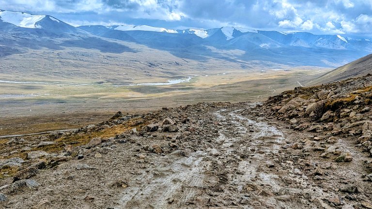 This landscape photo, likely from a bikepacking trip, depicts a rugged, muddy, and rocky trail winding into the foreground. In the midground, a wide valley stretches out, with a winding river visible, flanked by dry, grassy plains. In the background, majestic snow-capped mountains rise under a cloudy sky. The overall scene suggests a challenging yet scenic route, typical of remote high-altitude terrain.