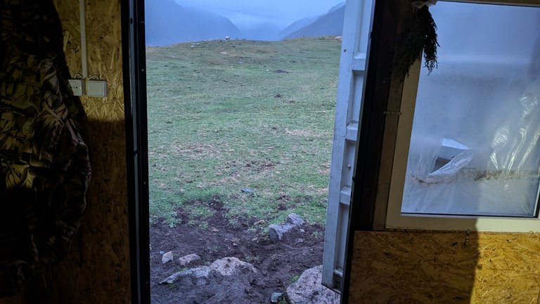 Looking out from inside a rustic shelter with walls made of OSB board, a doorway opens to a green, grassy hillside under a cloudy sky. In the foreground, the ground is muddy with visible rocks, suggesting an outdoor, rugged environment. Beyond the hillside, distant mountains are shrouded in mist. To the left inside the shelter, an electrical outlet and switch are visible, and a dark, patterned fabric hangs. To the right, a window with plastic sheeting covering part of it reveals more of the cloudy sky.