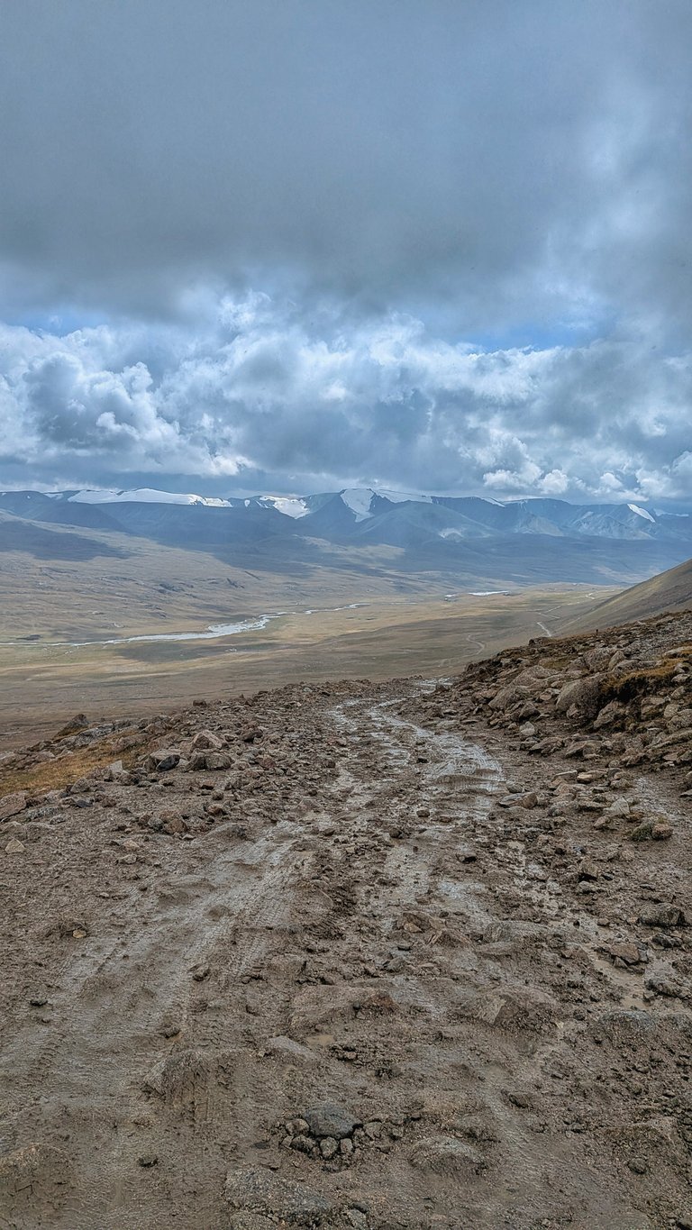 A vertical shot of a muddy and rocky trail leading down into a wide valley. In the foreground, the path is covered in dark, wet mud with visible tire tracks and scattered rocks. The trail winds slightly to the right, revealing a vast, light brown and green valley in the middle ground, through which a narrow river meanders. In the distance, a range of snow-capped mountains stretches horizontally across the horizon under a heavily clouded, grey sky, with hints of blue showing through in some areas. The scene depicts a remote, rugged landscape, typical of a challenging bikepacking route.
