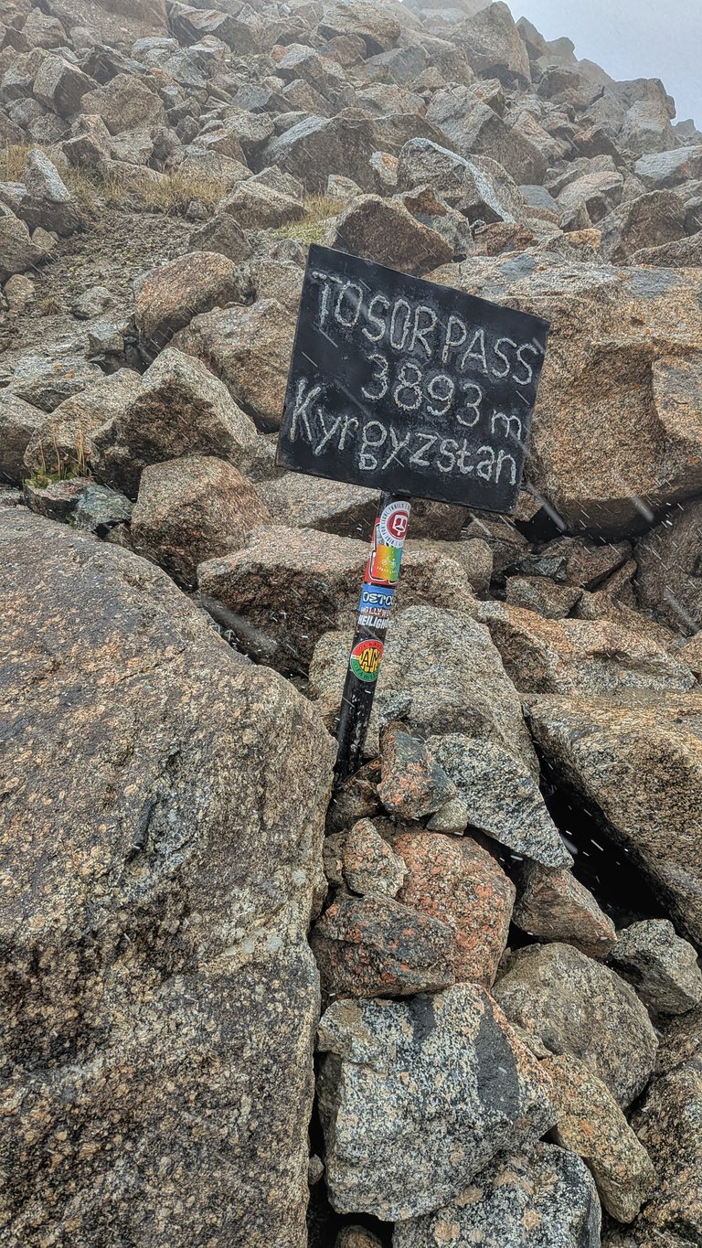 A close-up shot of a small, rustic sign planted among large, rugged rocks on what appears to be a mountain pass. The black sign, written in white chalk, reads "TOSOR PASS 3893 m Kyrgyzstan." The pole supporting the sign is covered in colorful stickers, including a rainbow flag design and others with text like "HOLLYWOOD HEILIGH." The ground is a mix of bare rock and sparse patches of dry grass, and flakes of snow or rain are visible in the air, suggesting a cold, possibly high-altitude environment. The scene captures a moment from a challenging bikepacking adventure in the mountains of Kyrgyzstan.