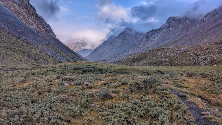 A wide shot of a rugged mountain valley, likely taken during a bikepacking trip. The foreground shows a patchy green and brown field with low-lying shrubs and a faint dirt path on the right. Steep, rocky mountains rise on both sides of the valley, their slopes covered in scree and sparse vegetation. In the distance, more peaks are visible, partially obscured by heavy, low-hanging clouds that dominate the sky, creating a moody atmosphere.