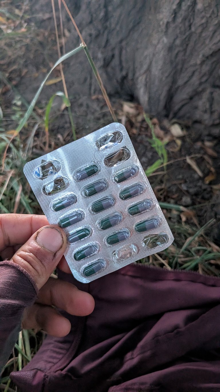 Levá ruka osoby s tmavým, možná špinavým palcem drží stříbrný blistr s pilulkami na pozadí řídké zelené trávy, tmavě hnědé země a texturovaného, tmavě šedého povrchu (pravděpodobně kmen stromu nebo kámen). Blistr obsahuje dvoutónové kapsle, zelené na jednom konci a světle šedé na druhém, s několika prázdnými místy naznačujícími, že některé pilulky byly užity. Osoba má na paži tmavě červenohnědý rukáv, který ladí s látkou viditelnou vpravo dole. Celkový dojem je venkovní prostředí, potenciálně na bikepackingovém výletě, se zaměřením na léky nebo doplňky stravy.