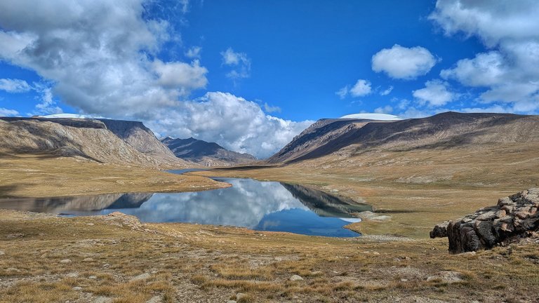 Široký panoramatický záběr krajiny ukazuje klidné jezero, ve kterém se odráží jasně modrá obloha s roztroušenými bílými mraky. Jezero je zasazeno mezi dvě drsné, hnědošedé hory, které se táhnou přes obzor. Hory jsou většinou pusté, s trsy suché, zlato-hnědé trávy v popředí a kolem okrajů jezera. Výrazná bílá, ploše zakončená plocha, pravděpodobně sníh nebo ledovec, je viditelná na vrcholu hory vpravo. V pravém dolním rohu vyčnívá výrazný shluk hnědých kamenů, dodávající scéně texturu. Celkový dojem je dojem vzdálené, vysokohorské divočiny, typické pro náročnou bikepackingovou trasu.