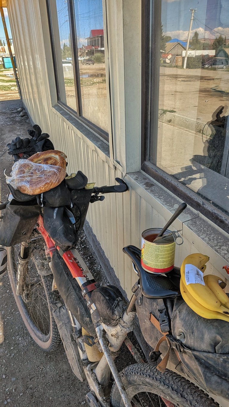 Špinavé bikepackingové kolo je opřené o světlou vlnitou plechovou stěnu s okny. Na řídítkách je připečený kulatý chléb připomínající pečivo, zabalený v plastu. Na zadním nosiči kola leží plechovka se lžící a trs banánů, což naznačuje zastávku na jídlo během dlouhé cesty. Okna odrážejí venkovskou scénu s domy, stromy a elektrickými dráty, což naznačuje venkovní, pravděpodobně odlehlé prostředí. Stav a naložení kola silně naznačují, že je uprostřed bikepackingového dobrodružství.