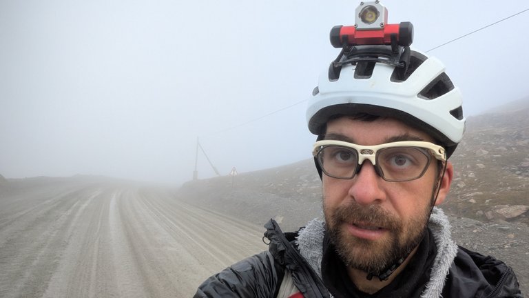 Muž s bílou cyklistickou přilbou s čelovkou a brýlemi s čirými obroučkami si dělá selfie a dívá se mírně nahoru. Má plnovous a na sobě černou bundu přes šedou chlupatou vrstvu. Za ním se do dálky táhne široká, blátivá polní cesta, zastřená hustou mlhou. Viditelnost je špatná, skrz mlhu jsou viditelné jen slabé obrysy kopců a vzdálená trojúhelníková dopravní značka.