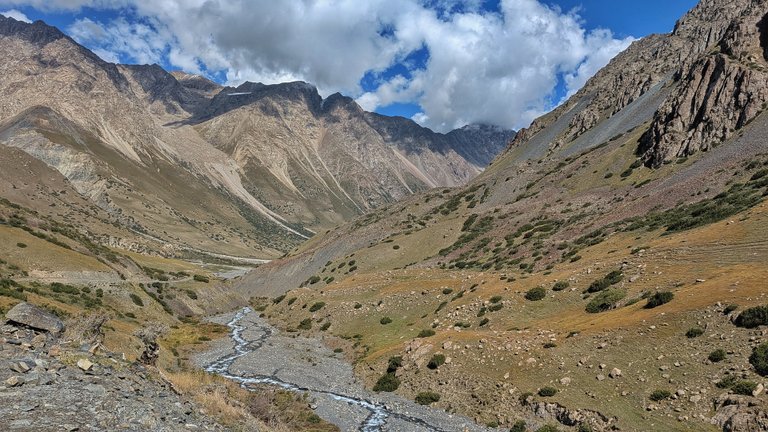 Širokoúhlá krajinářská fotografie zachycuje hluboké údolí v hornaté oblasti pod částečně zataženou modrou oblohou. Klikaté, kamenité koryto řeky s malým potokem protékajícím jím si razí cestu středem údolí a vede oko k vzdáleným horám. Svahy údolí jsou pokryty řídkou, zlato-hnědou a zelenou vegetací s roztroušenými kameny, což naznačuje náročný terén často potkáváný při bikepackingových dobrodružstvích. Vysoké, drsné hory s různými skalními útvary a suťovými svahy se majestátně tyčí po obou stranách údolí, jejichž vrcholy v některých oblastech mizí v oblacích, což naznačuje odlehlou a vysokohorskou polohu.