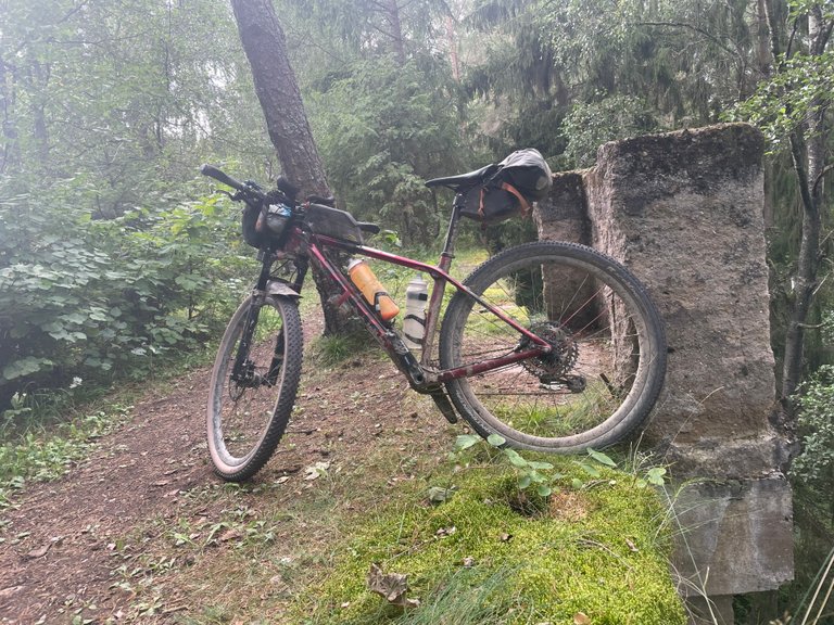 Z boku je vidět bikepackingové vybavení opřené o strom a starý kamenný sloup v hustém lese. Kolo je načervenalé horské kolo s předním odpružením, rámovou taškou, taškou na řídítka a podsedlovou taškou, což naznačuje, že je vybaveno na cestu. Na rámu je připevněna oranžová láhev na vodu. Cesta pod kolem je z hlíny a kořenů, obklopená zeleným podrostem a mechem. V pozadí je vidět mnoho stromů se světle zeleným listím, což naznačuje živé přírodní prostředí.
